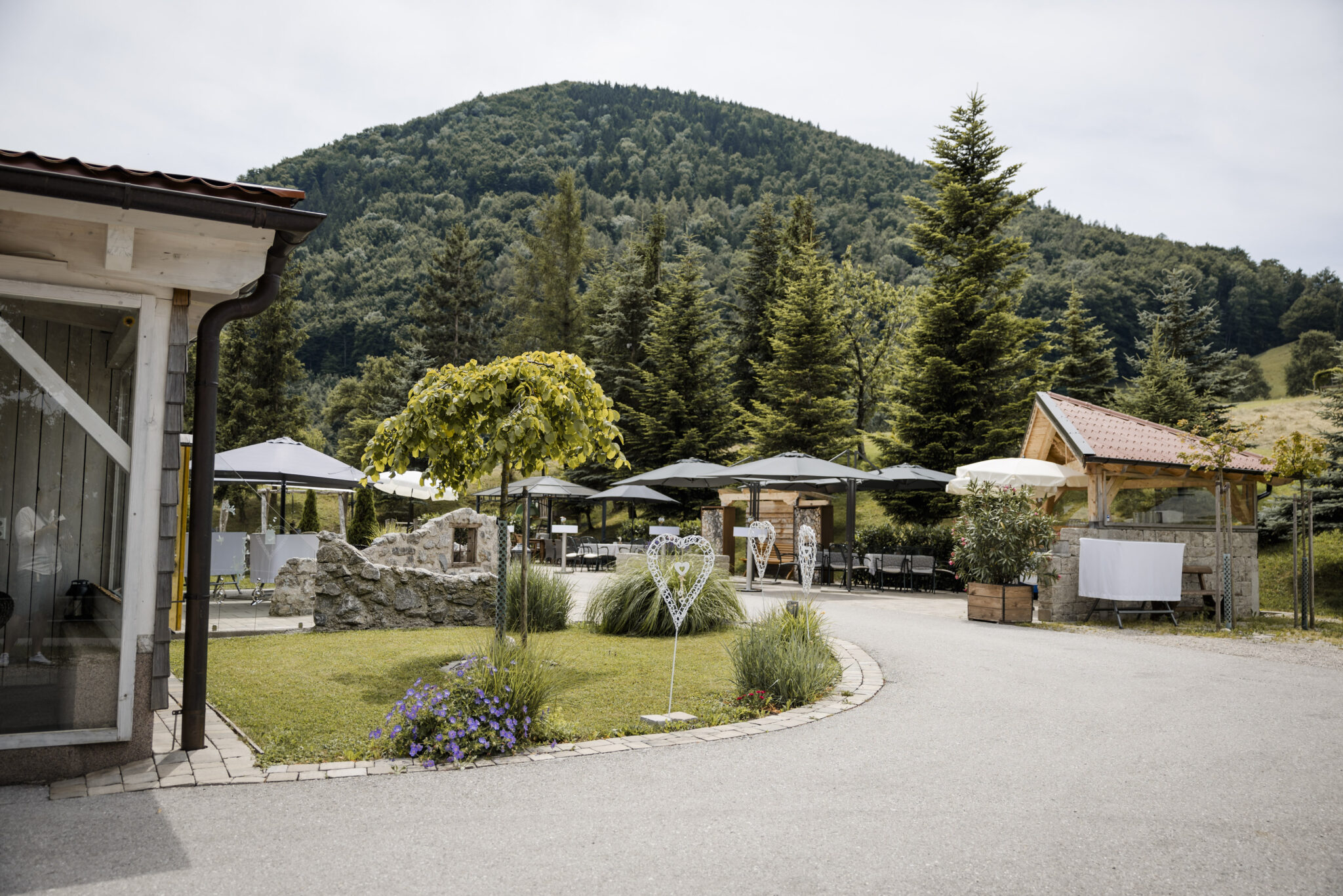 Eine Terrasse im Freien mit Tischen und Sonnenschirmen in der Nähe eines grünen, bewaldeten Berges unter teilweise bewölktem Himmel – ein idealer Ort für eine Hochzeit oder eine entspannte Firmenfeier, mit Übernachtungsmöglichkeiten in der Nähe.
