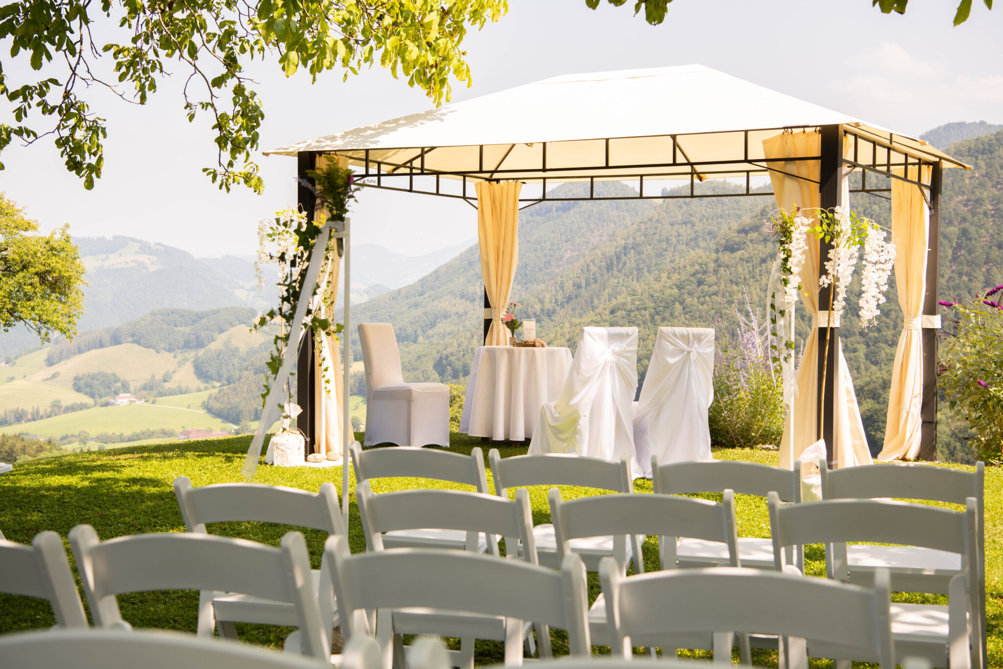Hochzeitslocation im Freien mit weißen Stühlen und einem geschmückten Pavillon mit Blick auf grüne Hügel und Berge – ideal für Privatfeiern oder auch Firmenfeiern. Optionale Übernachtungsmöglichkeiten für Gäste sind verfügbar.