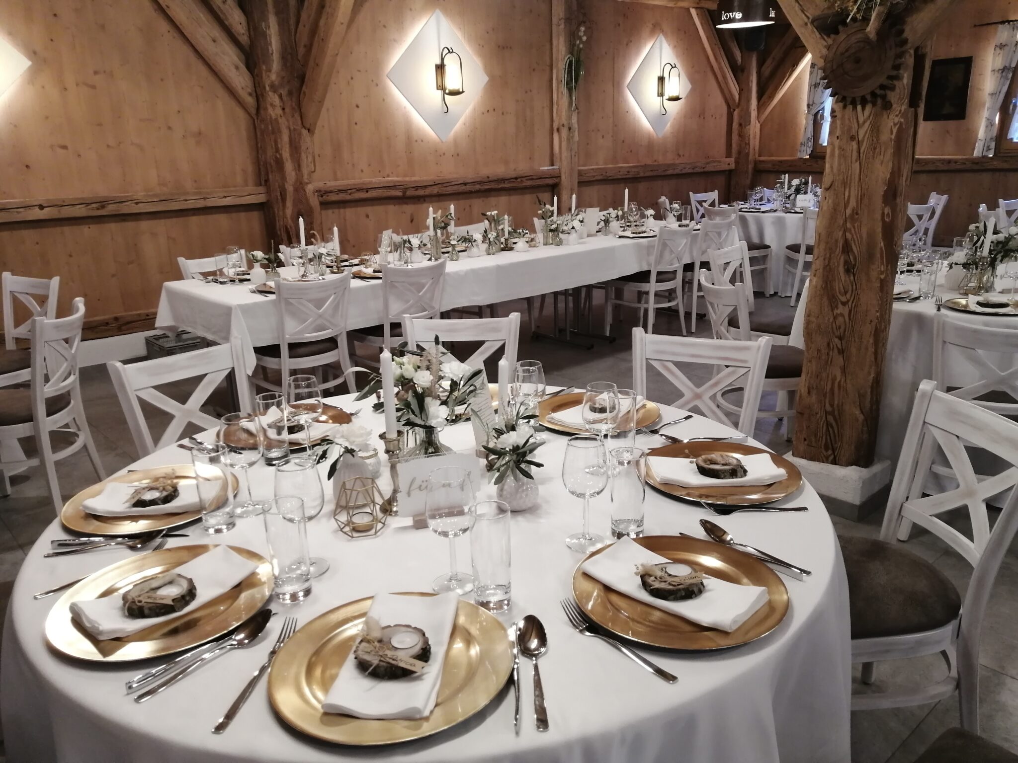 Runde und rechteckige Tische, die für eine feierliche Veranstaltung mit weißen Tischdecken, goldenen Tellern und Blumengestecken gedeckt sind, schaffen in dieser atemberaubenden Veranstaltungslocation eine elegante Atmosphäre für eine Firmenfeier.