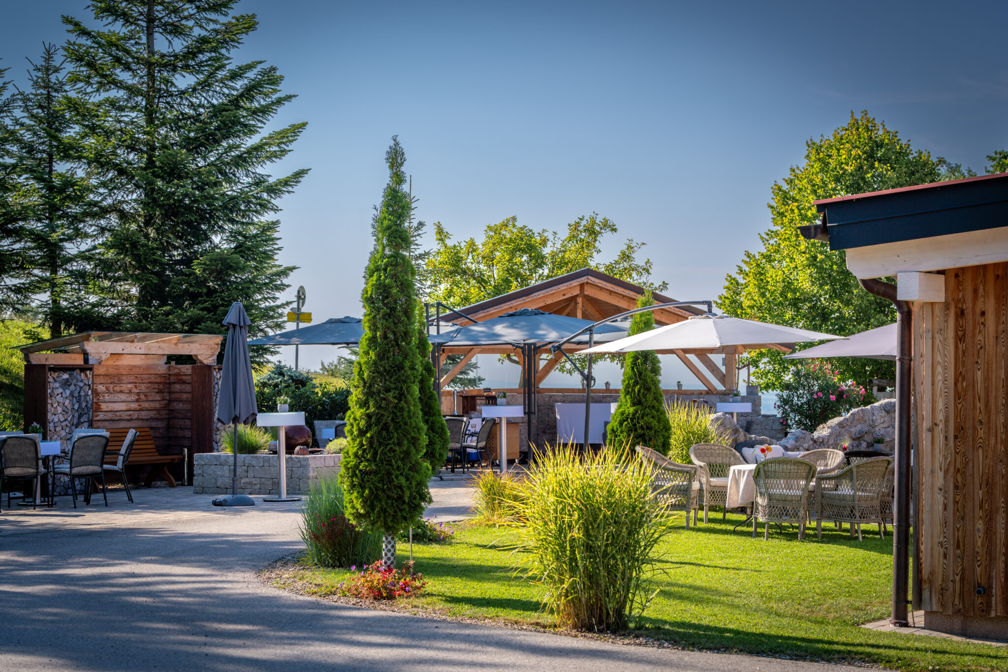 Eine Terrasse im Freien mit Tischen, Stühlen, Sonnenschirmen, viel Grün und Holzkonstruktionen unter strahlend blauem Himmel – ideal für Ihre Firmenfeier oder eine private Übernachtung.