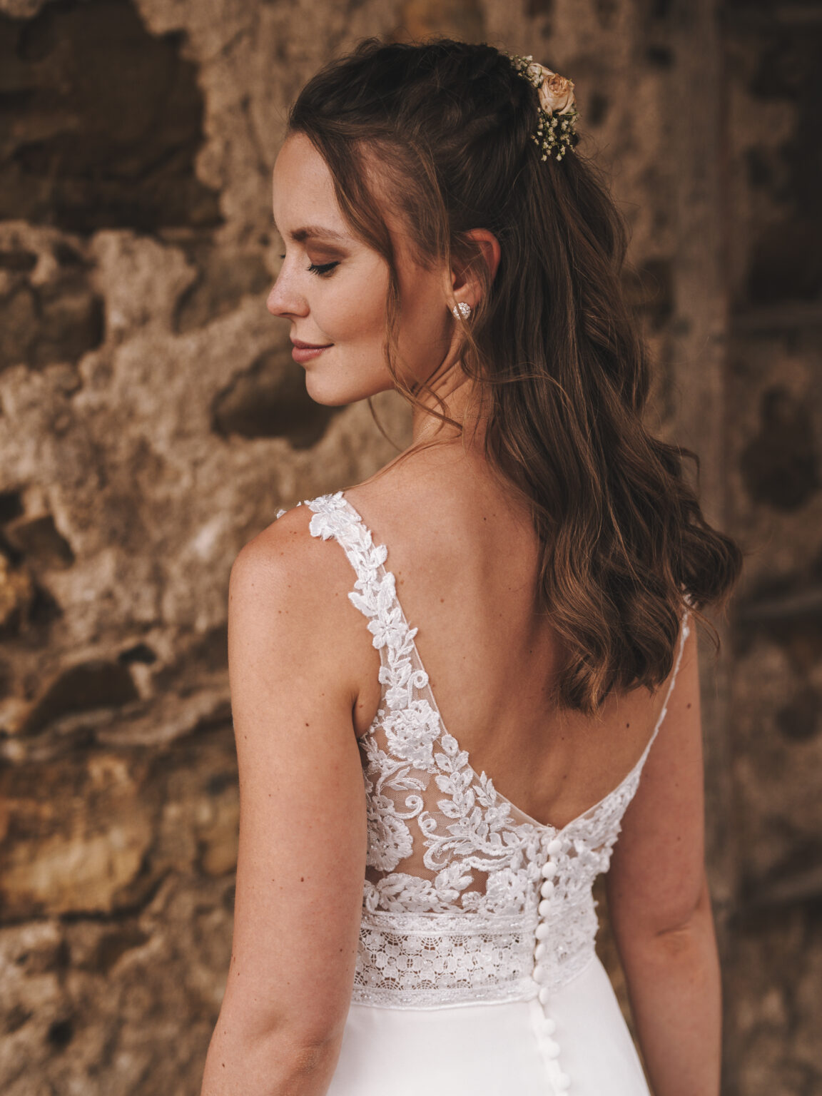Eine Braut in einem Spitzenhochzeitskleid steht mit dem Rücken zur Kamera und lächelt sanft vor einer rustikalen Steinmauer – ein eleganter Moment, der sich perfekt für jede Privatfeier oder Firmenfeier in intimem Rahmen eignet.