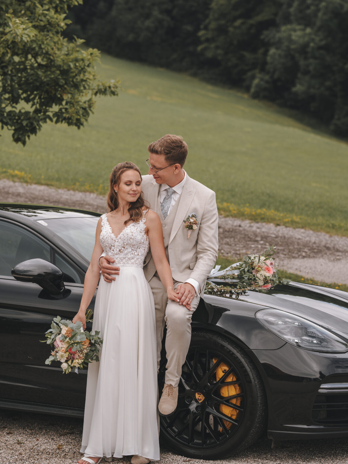 Braut und Bräutigam in Hochzeitskleidung posieren mit Blumensträußen vor einem schwarzen Auto, im Hintergrund ist Grün zu sehen – perfekt, um Erinnerungen an Ihre Privatfeier festzuhalten.
